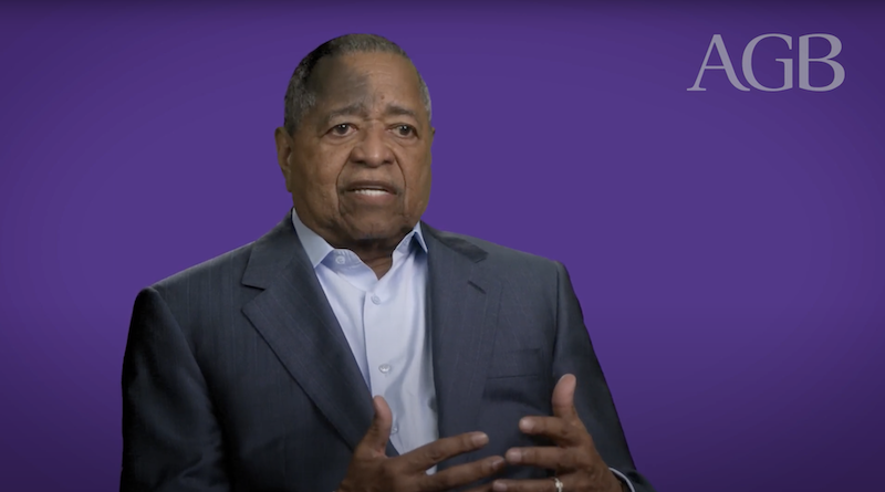 Man wearing a dark suit talking in front of a purple background with the letters A, G, B.